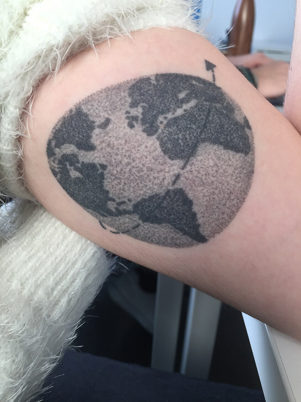 Tatuaje circular de la Tierra en un brazo, sombreado con puntos y acompañado por la silueta de un avión.