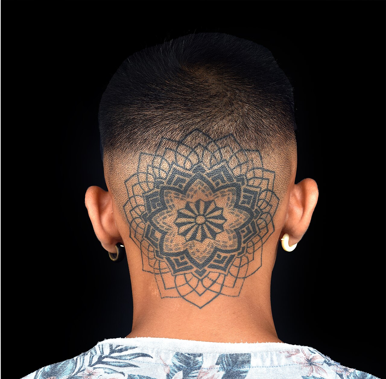 Mandala tatuado en la parte posterior de la cabeza, con contornos negros, puntos y espacios de piel sin tinta.